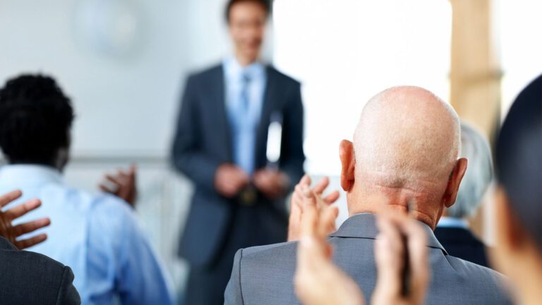 Person standing at front of room with audience applauding